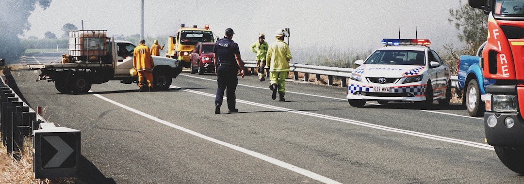 Image of a traffic incident on a two-lane road. A policeman and first responders stand assessing the scene.